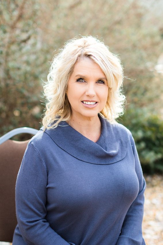 Professional headshot of person in blue cowl neck sweater against outdoor setting.