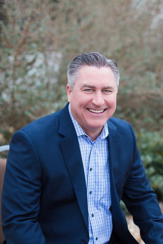 Professional headshot of person in navy suit and checkered shirt against outdoor setting.