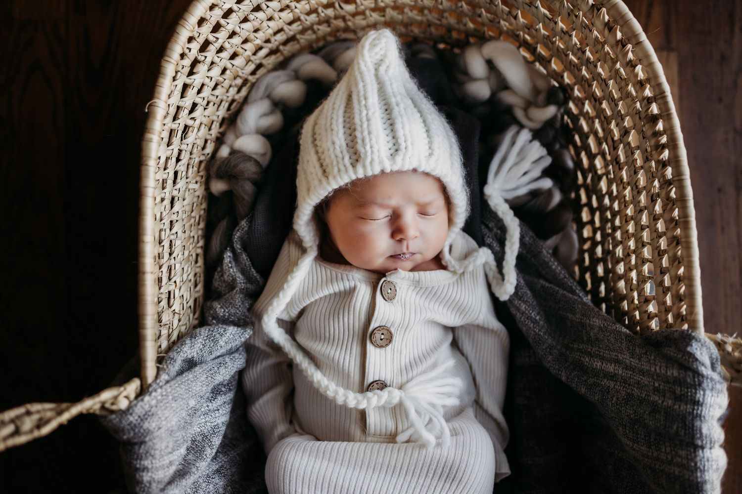 Baby in white knit outfit and bonnet lying in woven basket with blankets.
