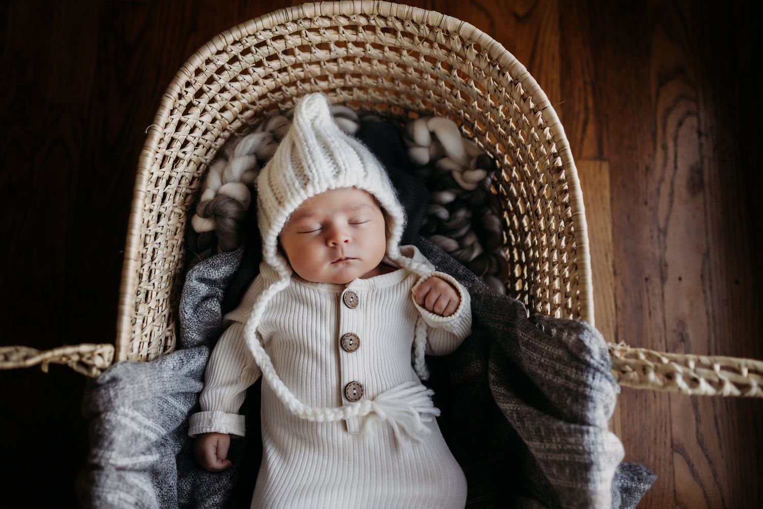 Baby wearing white knit clothing resting in woven basket with cozy grey lining.