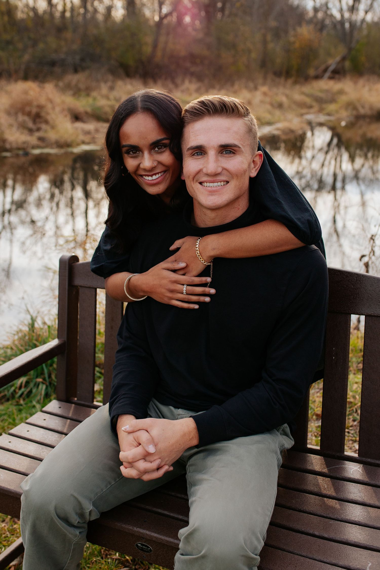Couple sitting close together on wooden bench by river in autumn setting.