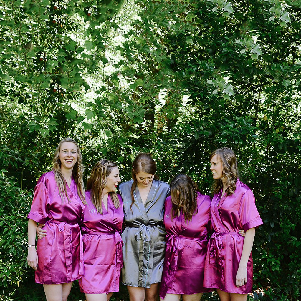 Bridal party wearing pink and grey satin robes posing together in front of green foliage.