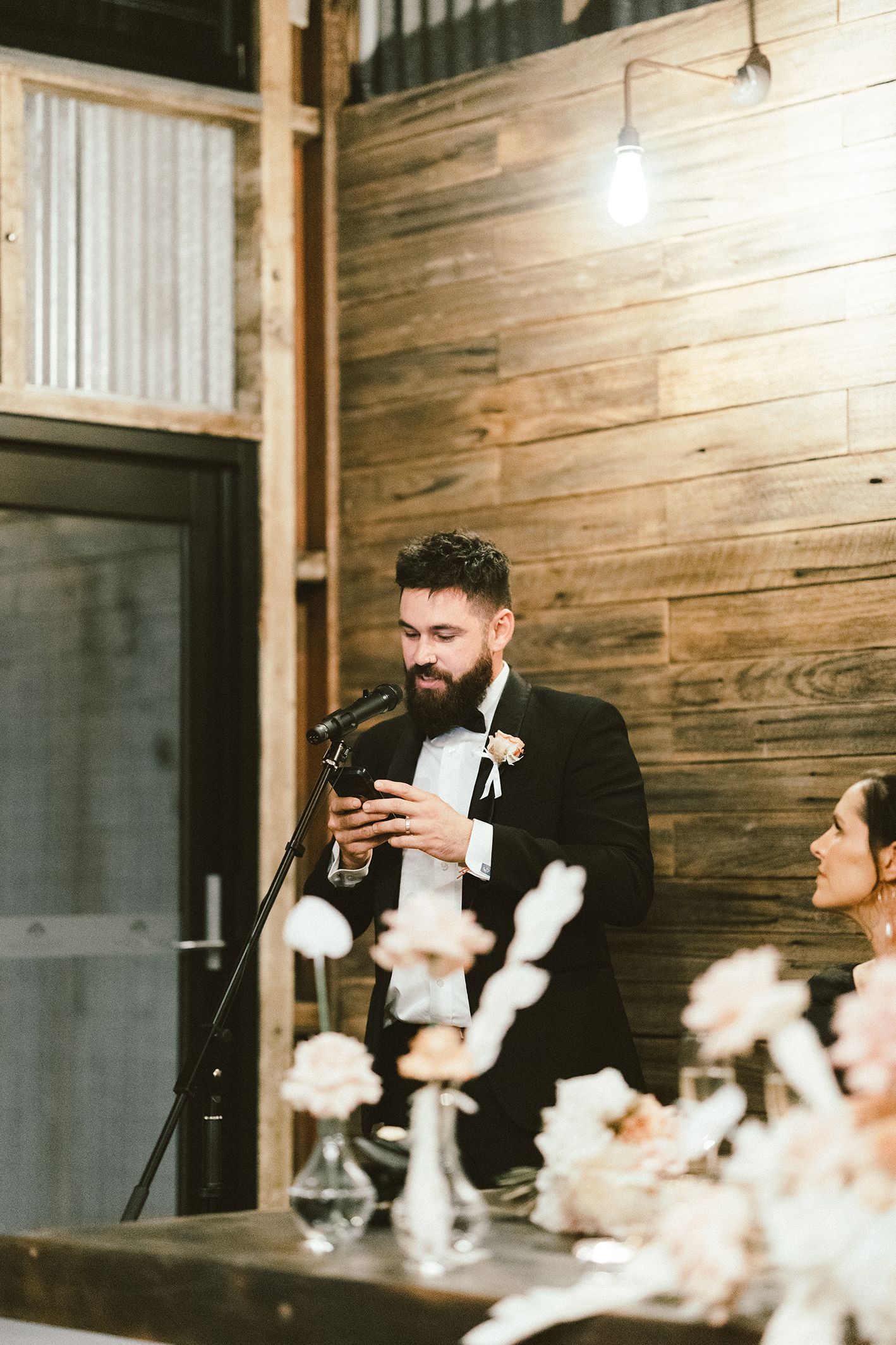 Best man giving speech at rustic wedding reception with wooden wall backdrop.