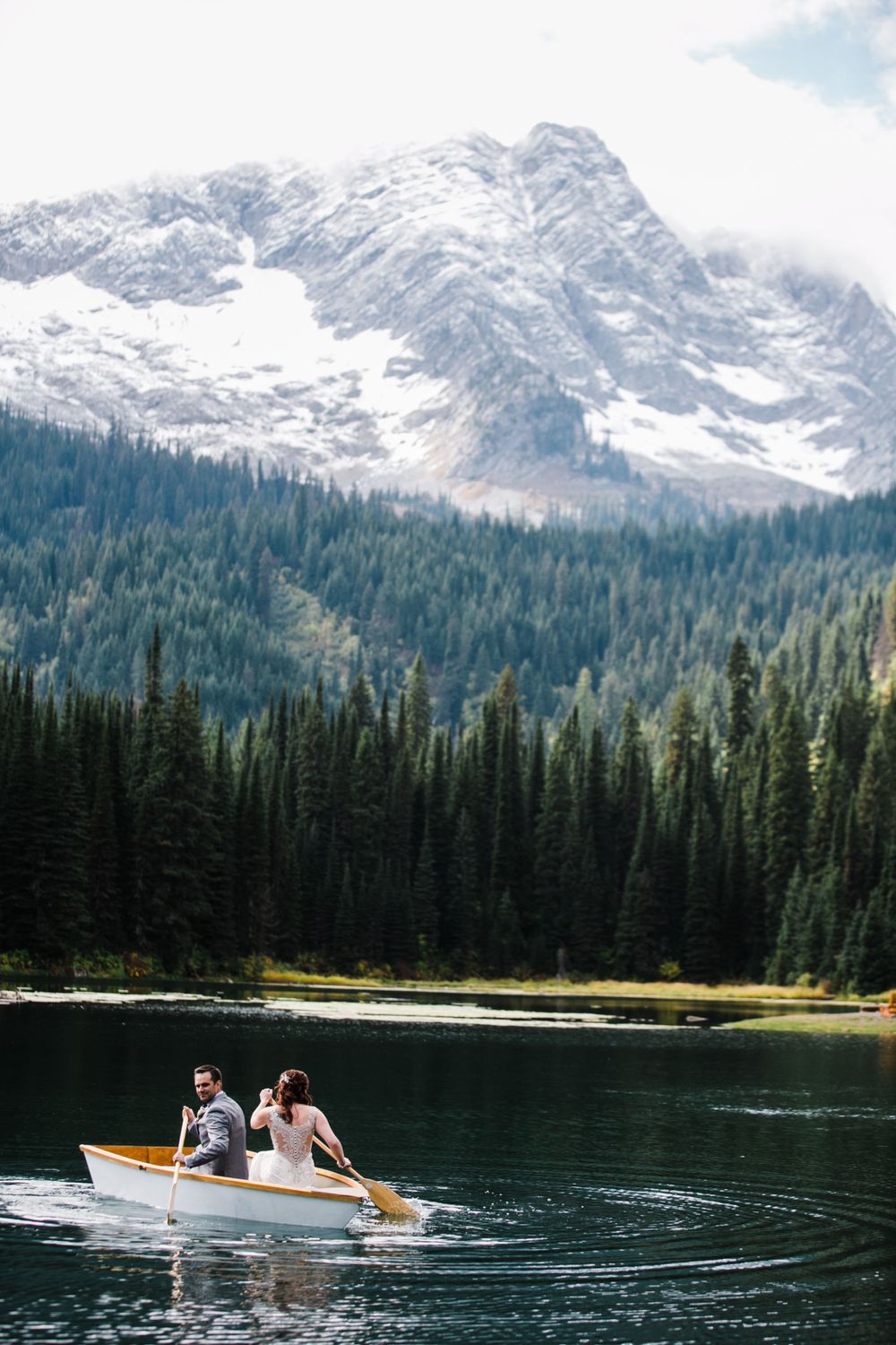 Wedding party rides in wooden boat across pristine mountain lake surrounded by evergreen forest.