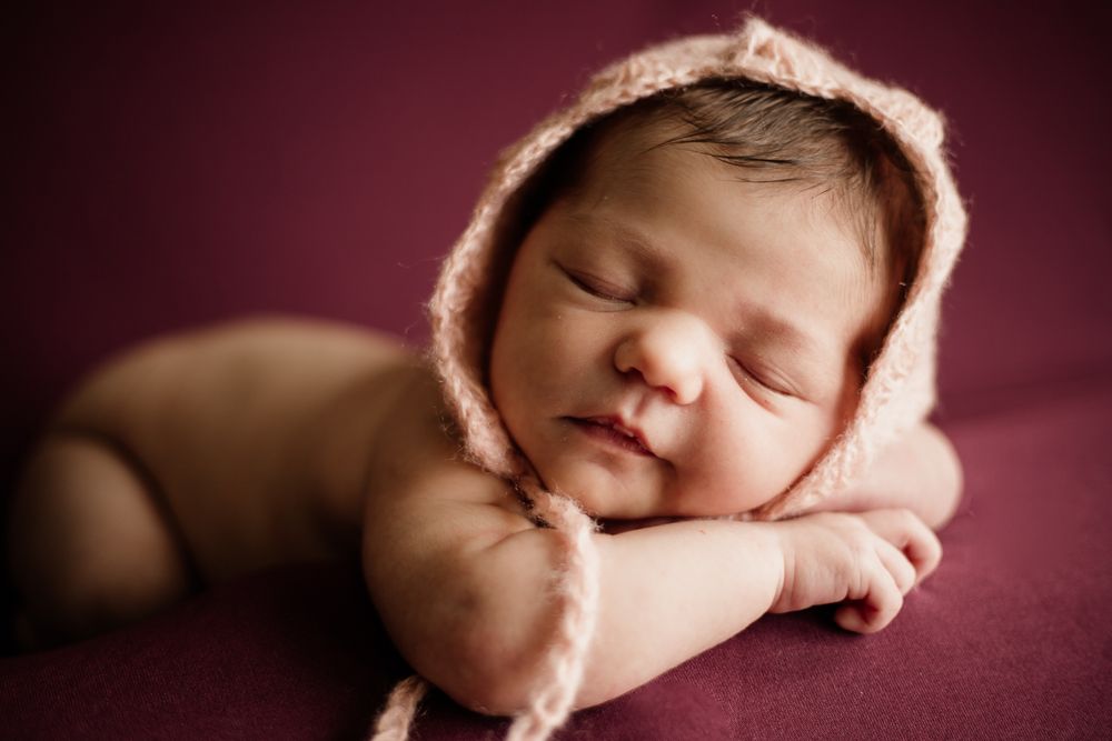 Close-up portrait of sleeping newborn with pink bonnet against burgundy background.