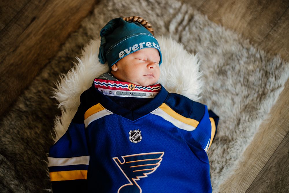 Newborn baby wearing St. Louis Blues hockey jersey sleeping on textured backdrop.