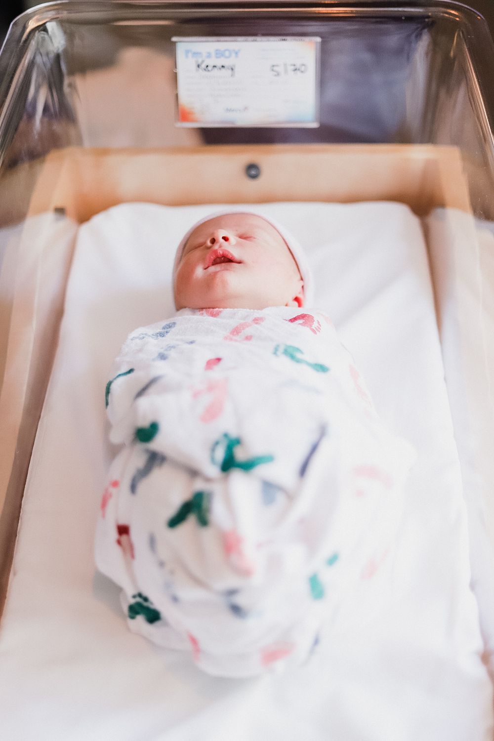 Newborn baby sleeping in hospital bassinet wrapped in patterned swaddle blanket.