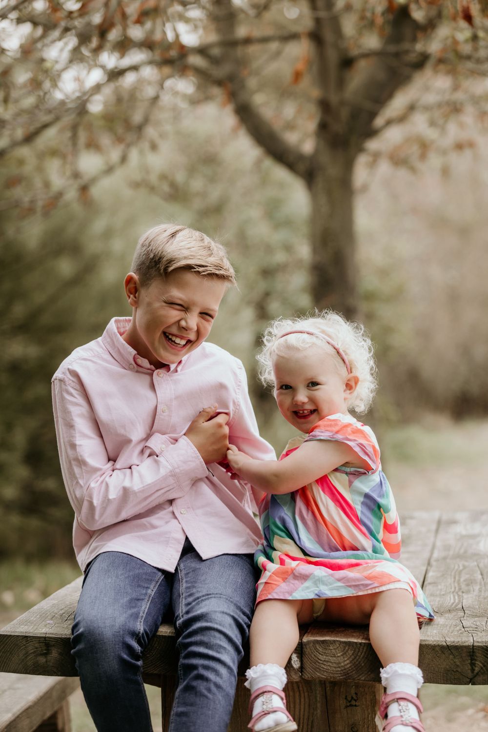 Two people share a joyful moment while sitting on a wooden bench in a park setting.