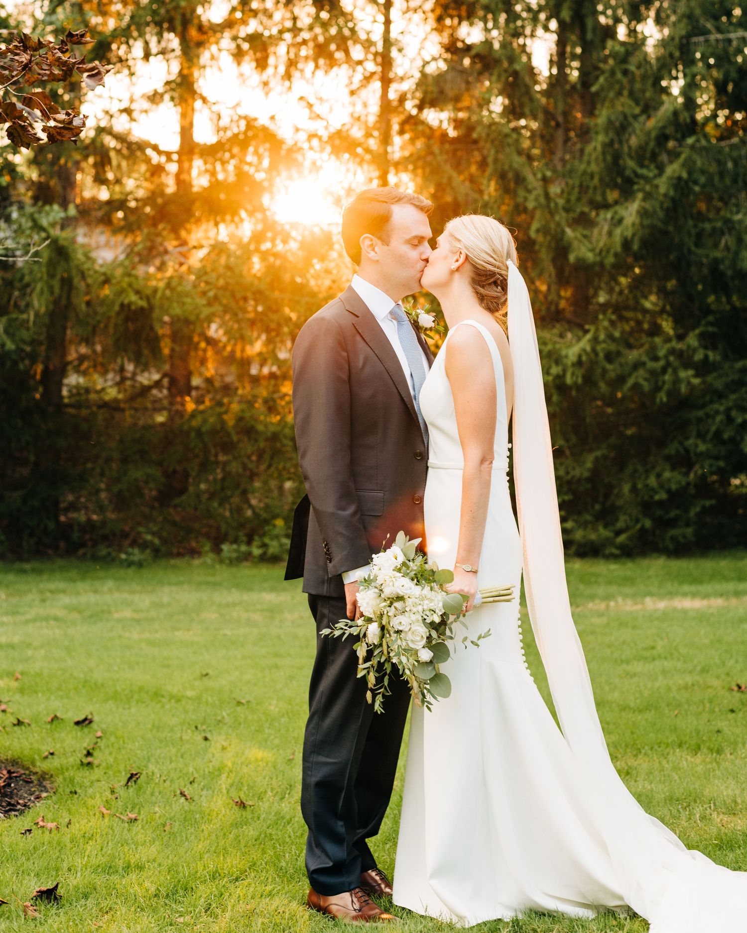 Romantic sunset wedding portrait with couple among trees holding white bouquet.