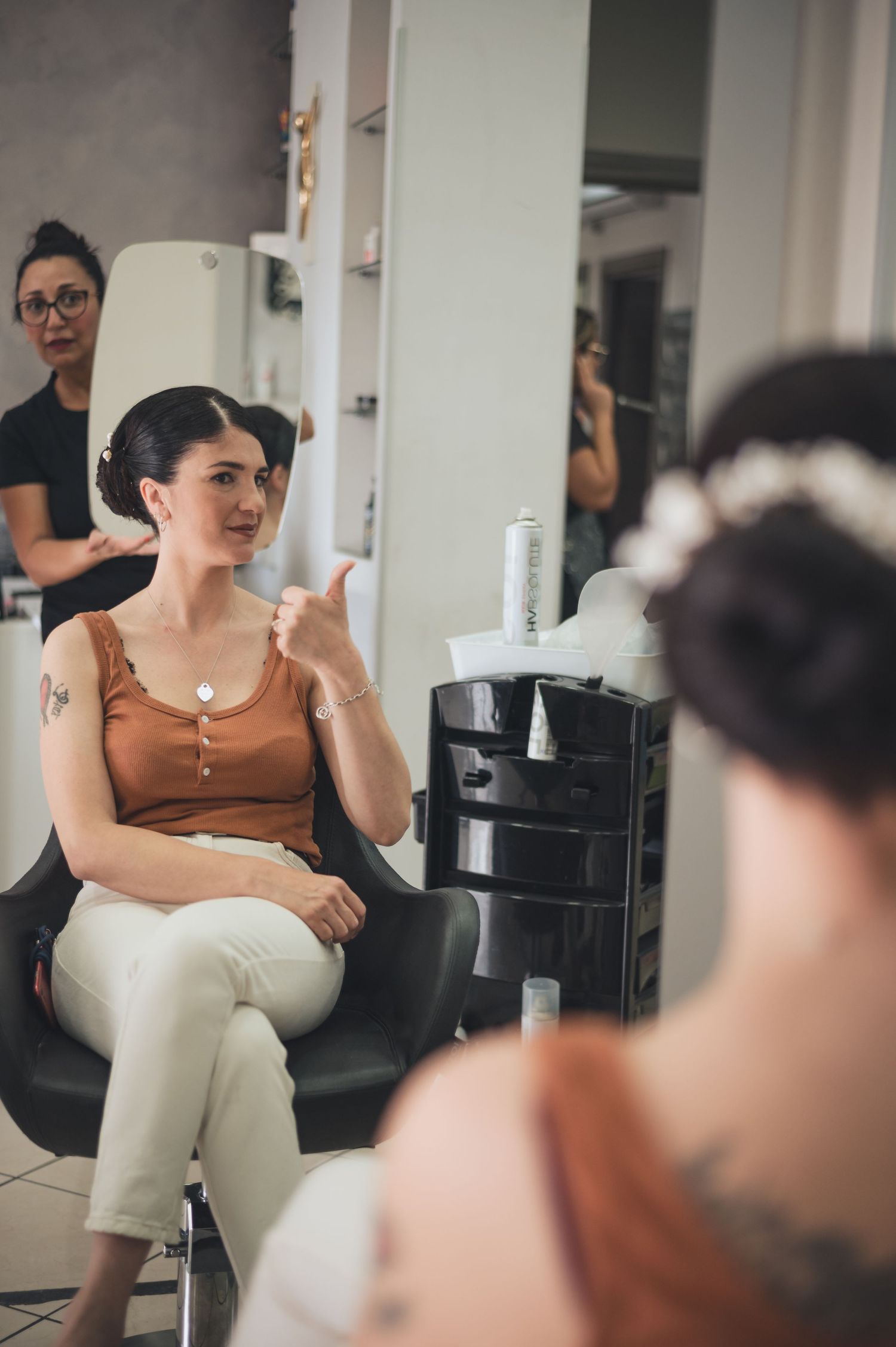 Client sits in salon chair wearing brown tank top and white pants during beauty appointment.