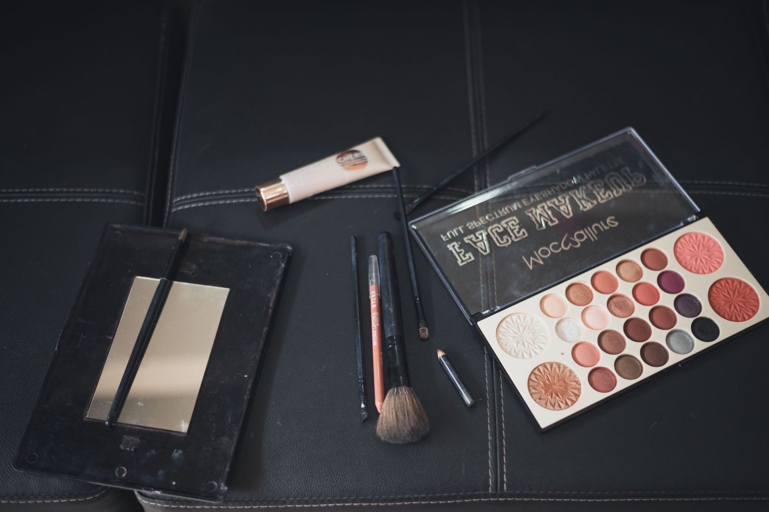 Flatlay of beauty products including eyeshadow palette, brush and mirror on dark surface.
