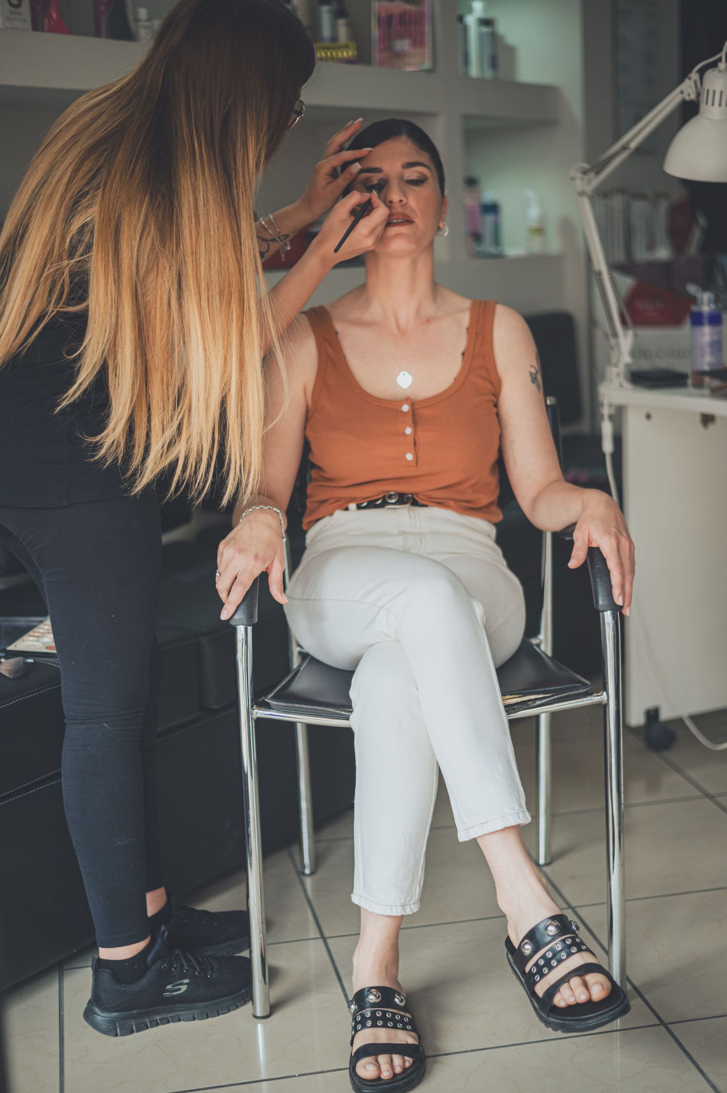 Beauty professional applies makeup to client wearing orange tank top in salon setting.