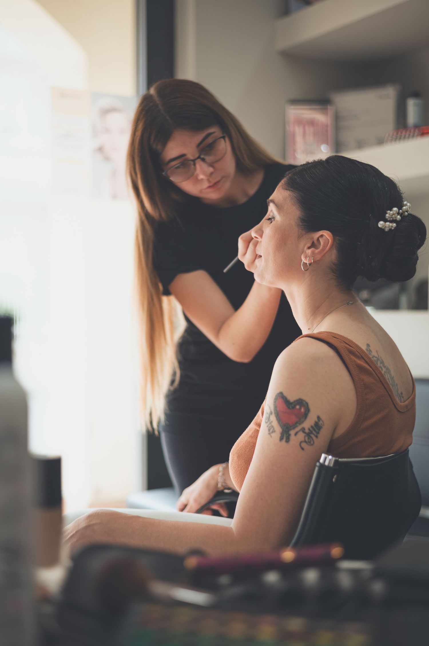 Makeup artist works on client's look in beauty salon with visible arm tattoo.