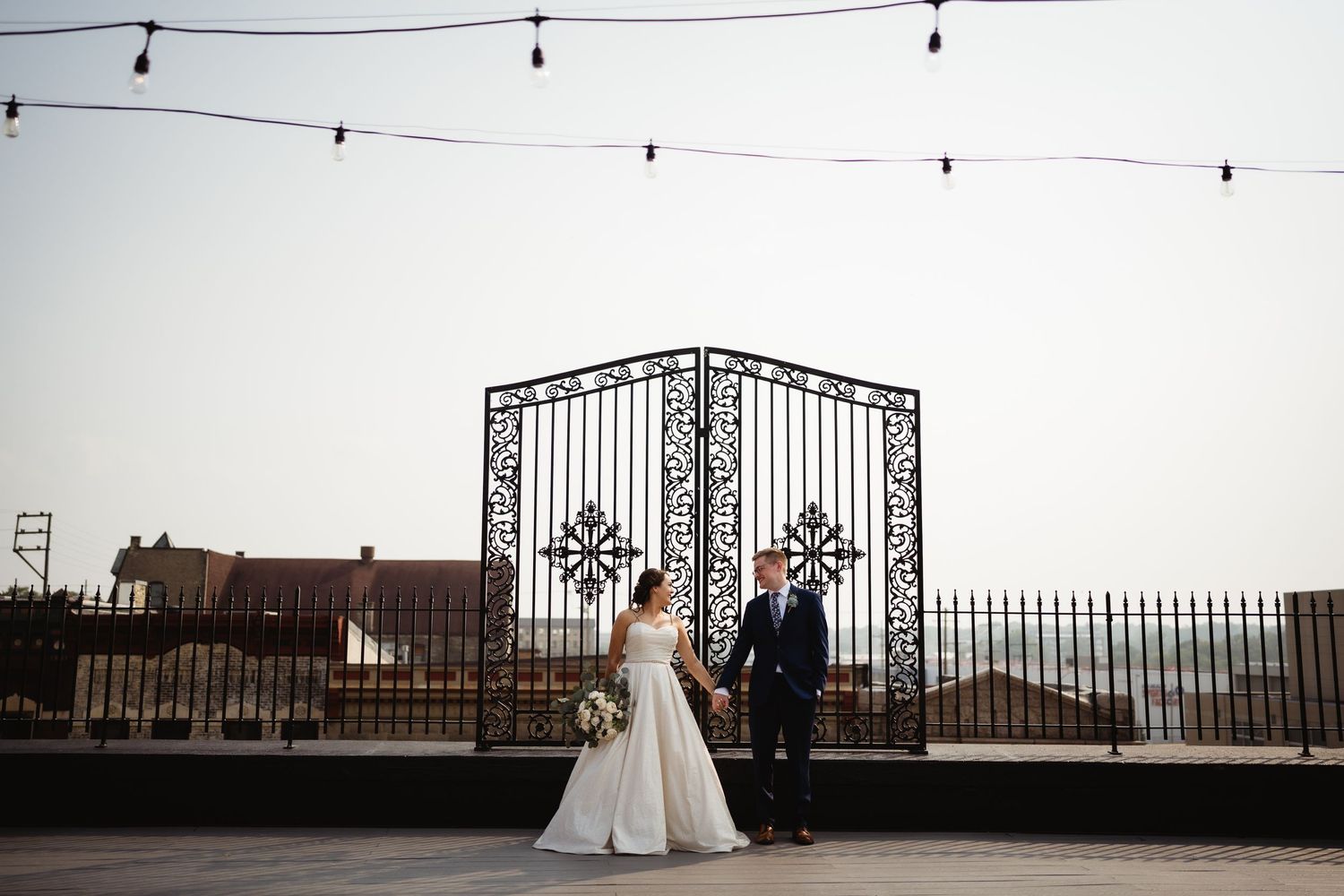 Wedding couple standing under string lights by ornate iron gates on a rooftop venue at dusk.