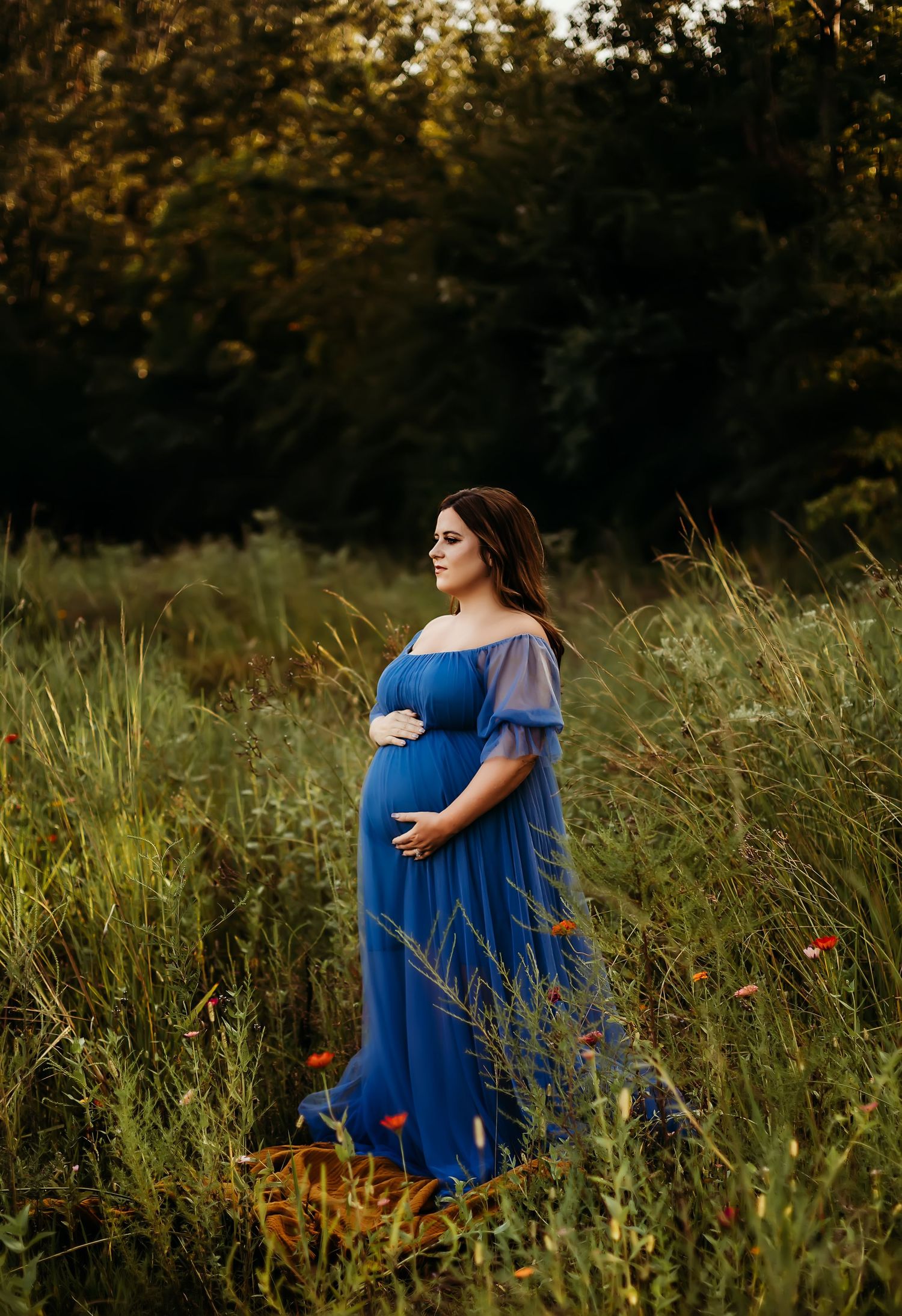 A pregnant woman in a flowing blue off-shoulder dress stands in a sunlit meadow with wildflowers.