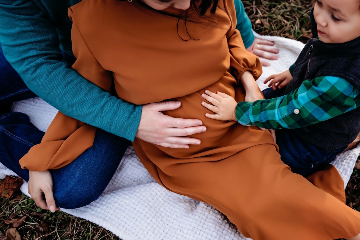 Close-up of hands touching pregnant belly in rust-colored dress on white blanket.