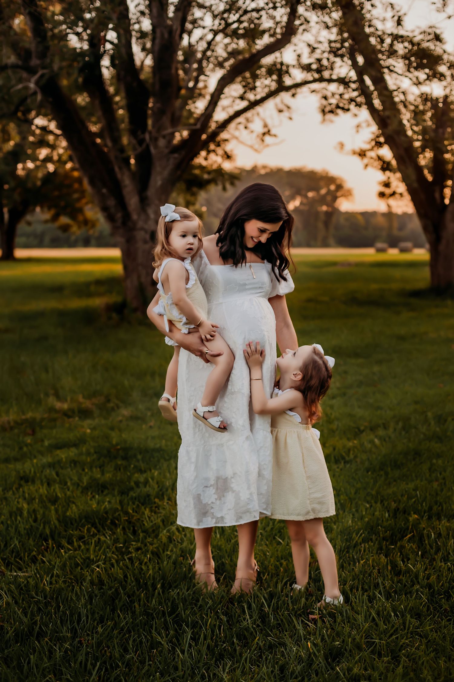 A tender outdoor portrait at sunset shows a pregnant woman with two young children in light dresses.