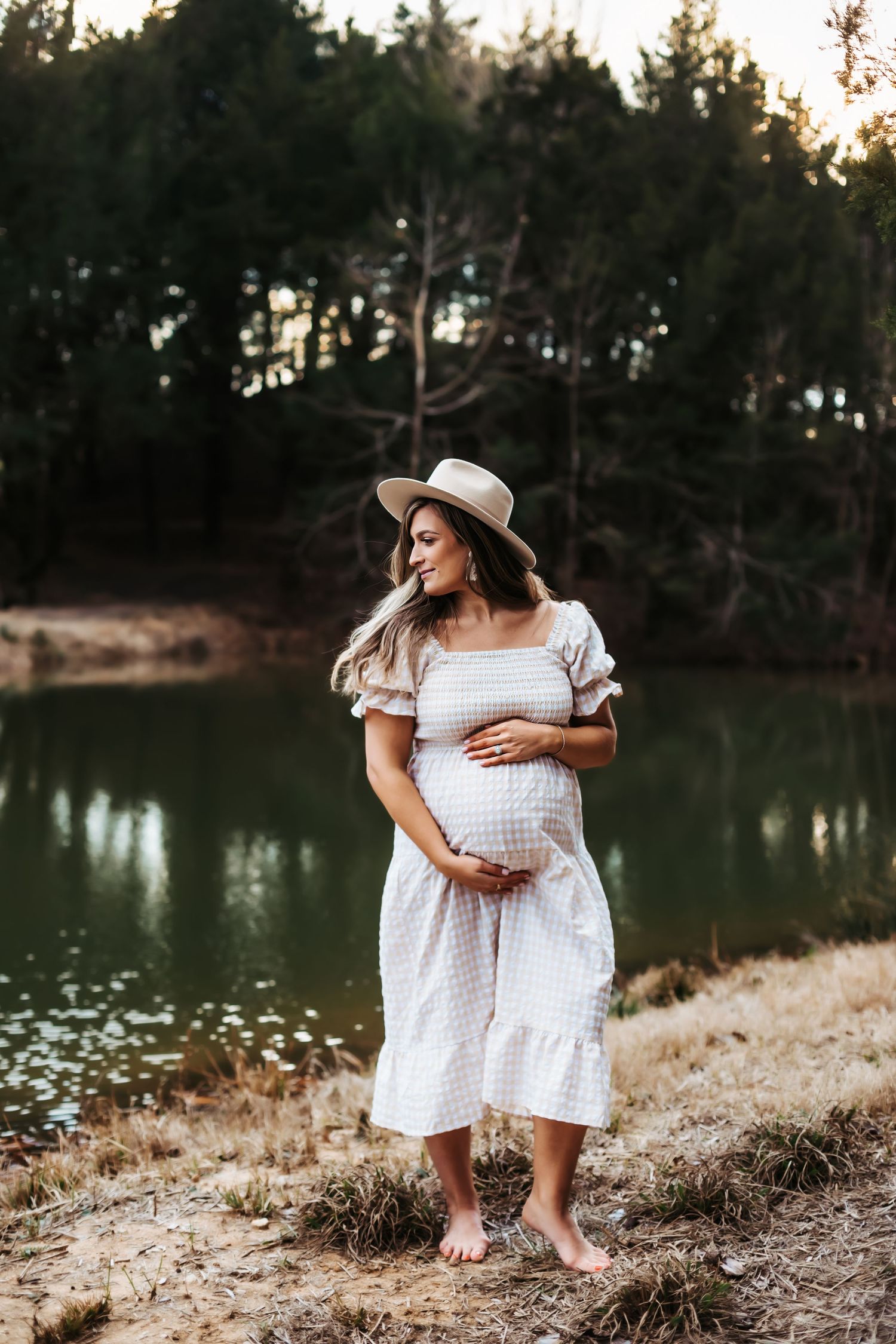 Expectant mother in white sundress and hat poses barefoot by serene lake at sunset.