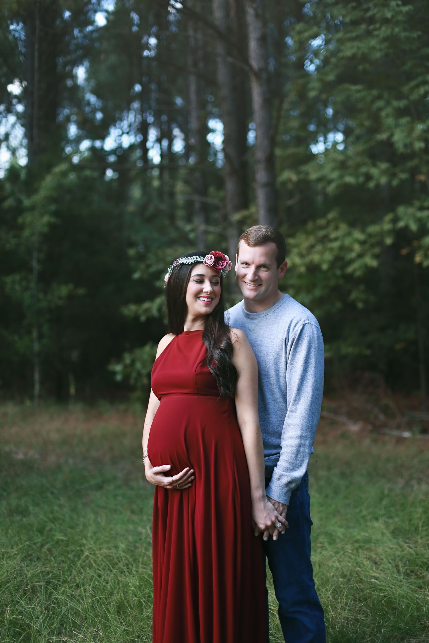 A couple poses in a forest setting with the woman wearing a flowing red maternity dress.