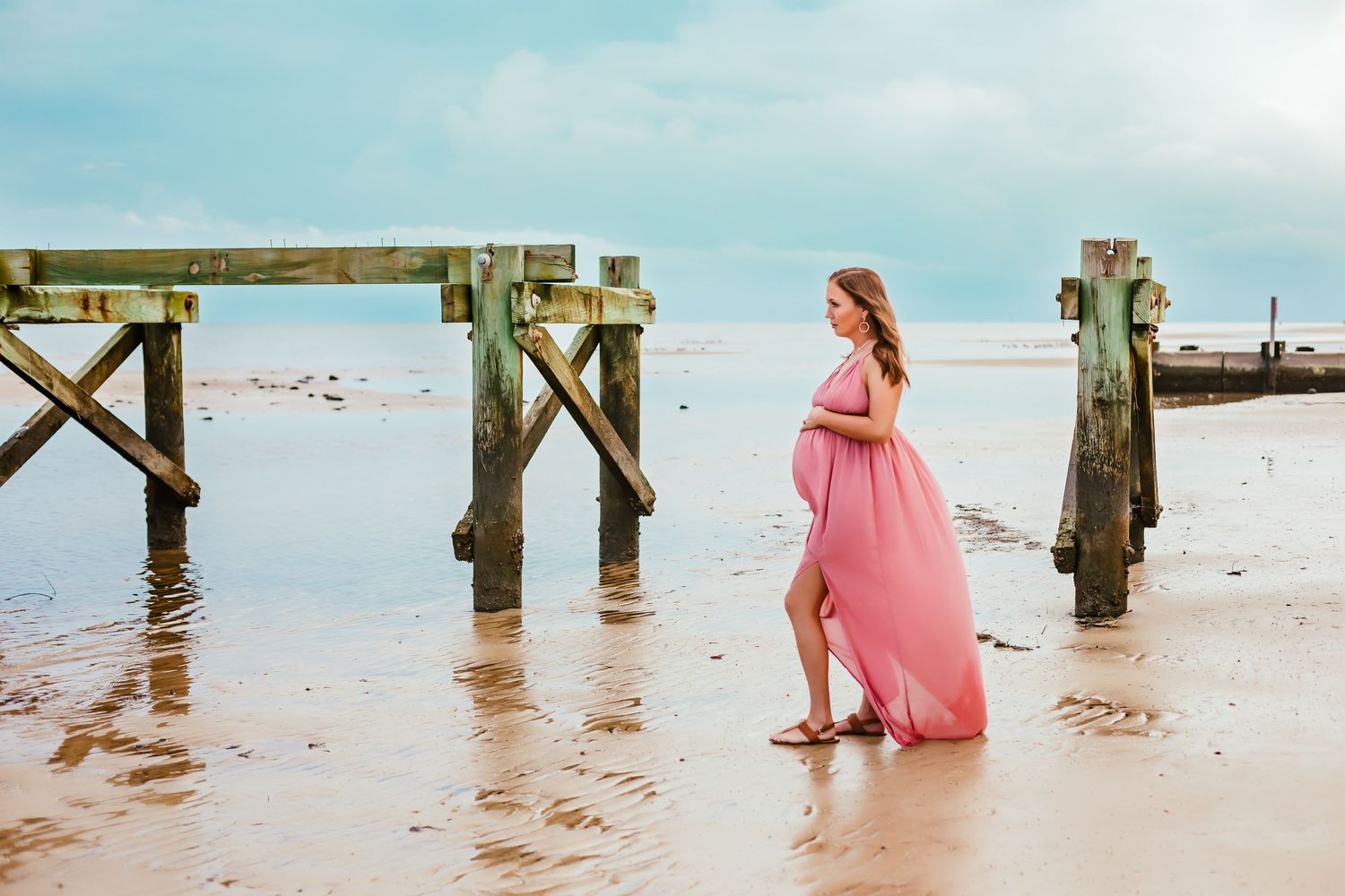 A flowing pink dress catches the breeze near wooden pier posts on a beach at sunset.