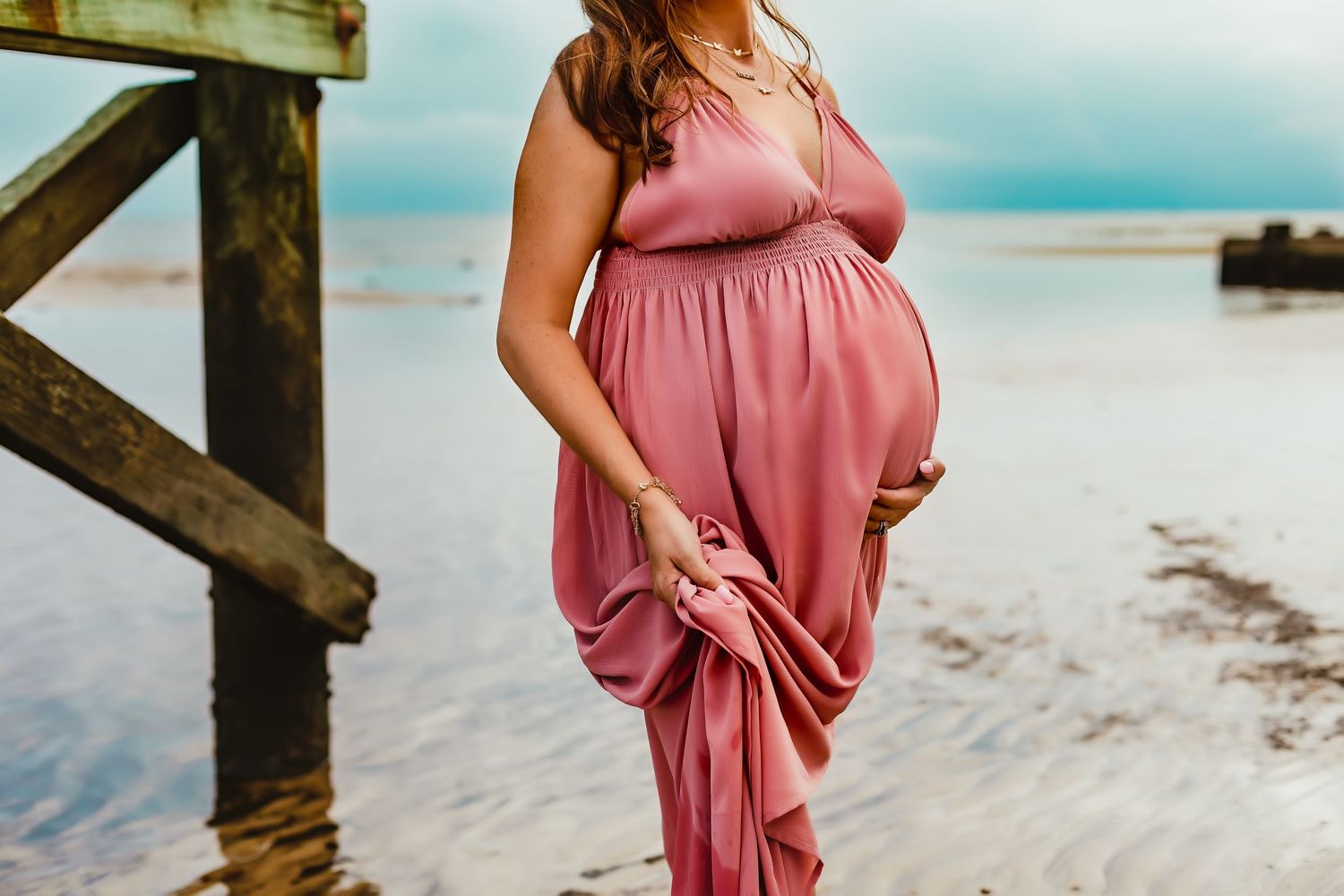 A pink maxi dress billows in the coastal breeze near weathered wooden pier posts.