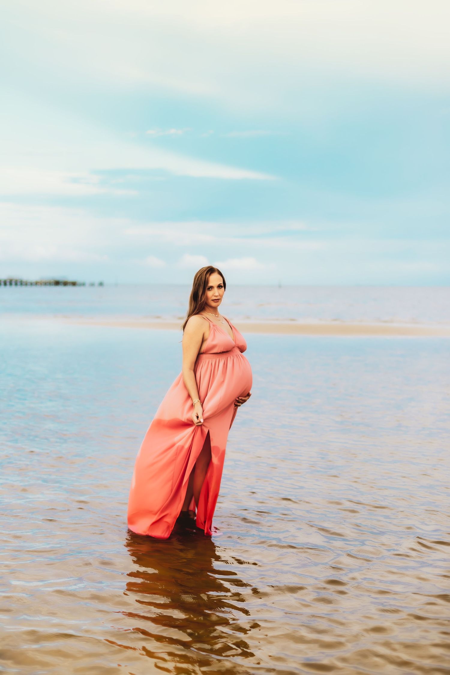 A coral floor-length dress flows gracefully in shallow ocean waters against a dreamy blue sky.