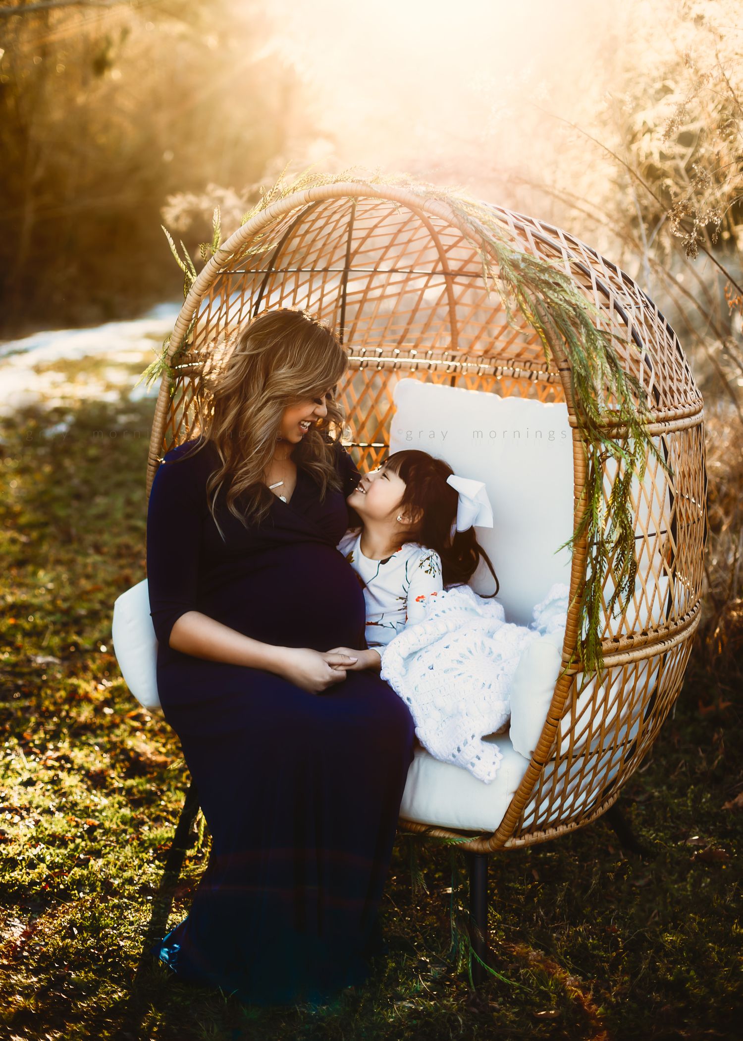 Intimate maternal moment captured in woven chair with natural backlight and greenery.