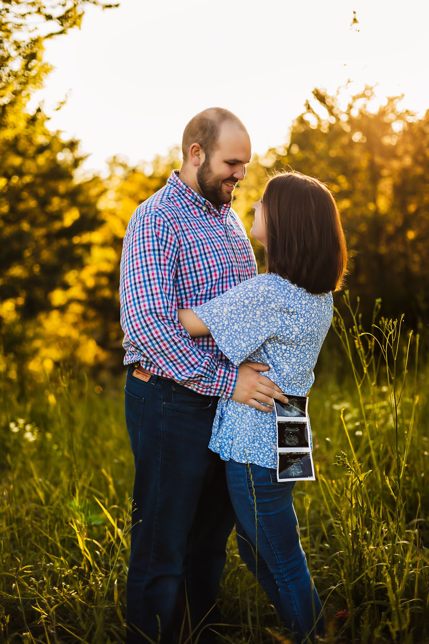 A couple embraces in golden sunset light while holding ultrasound photos.