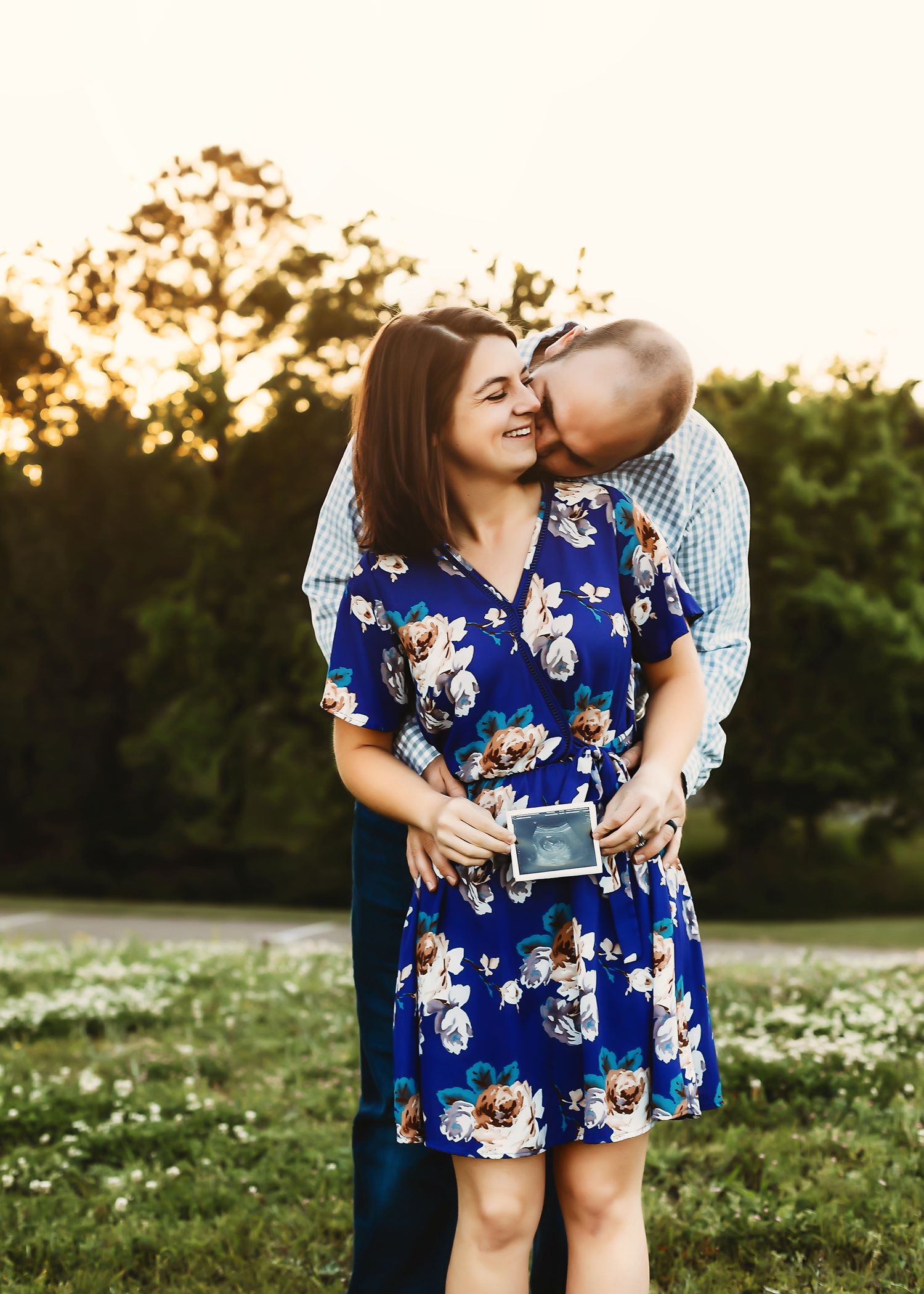 Couple sharing tender moment in sunlit field as they reveal ultrasound photo during pregnancy announcement.