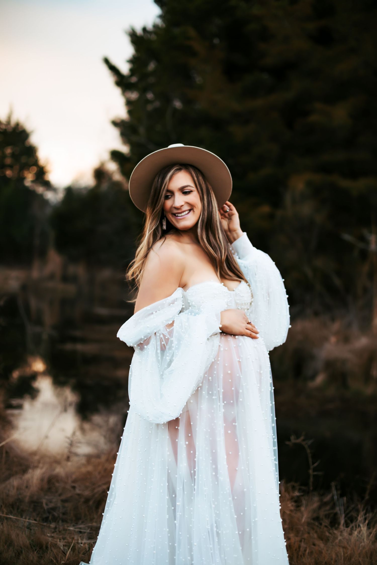 Bohemian maternity photo featuring white lace dress and wide-brimmed hat in nature setting.