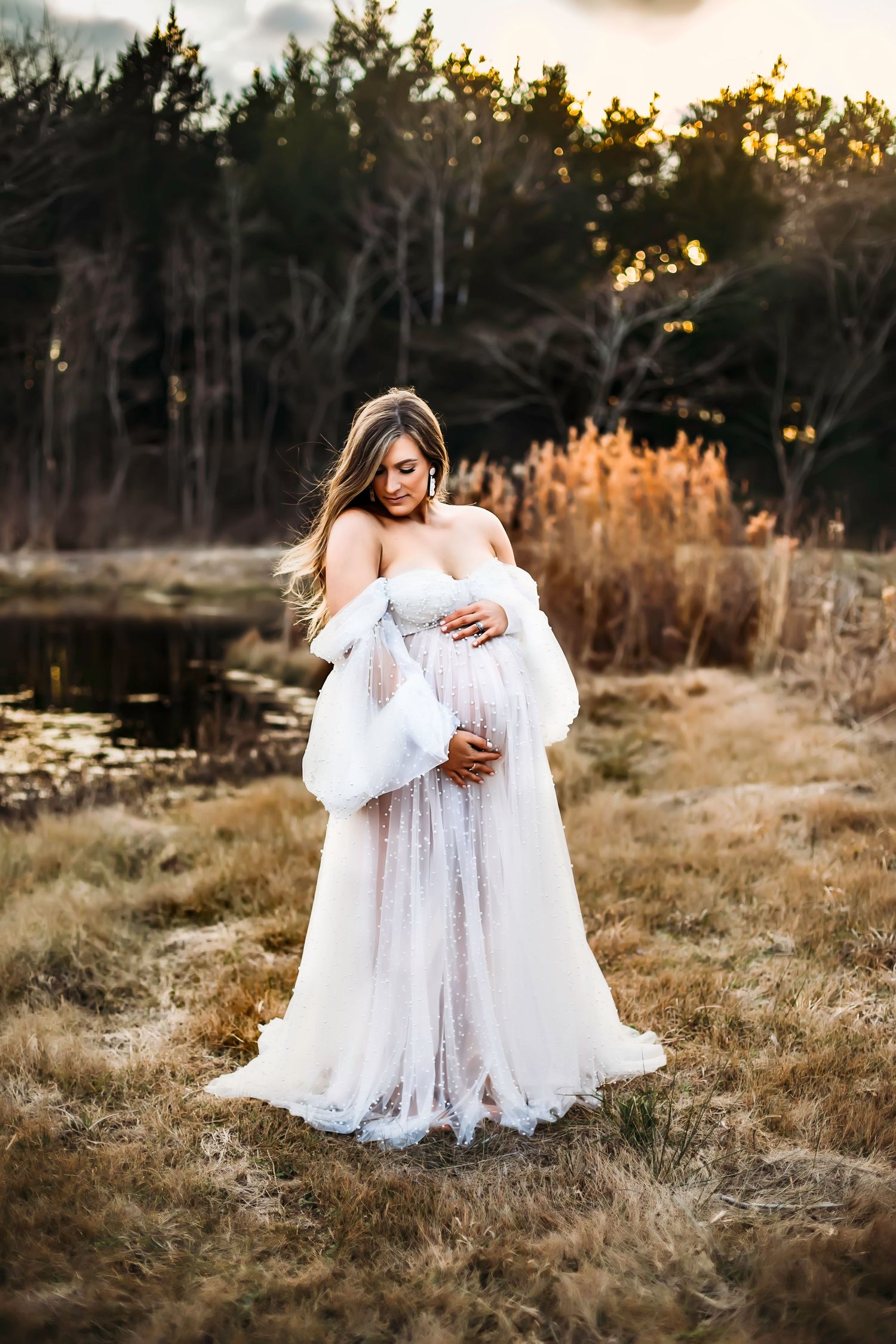 Ethereal maternity portrait in flowing white gown against golden sunset and lake backdrop.