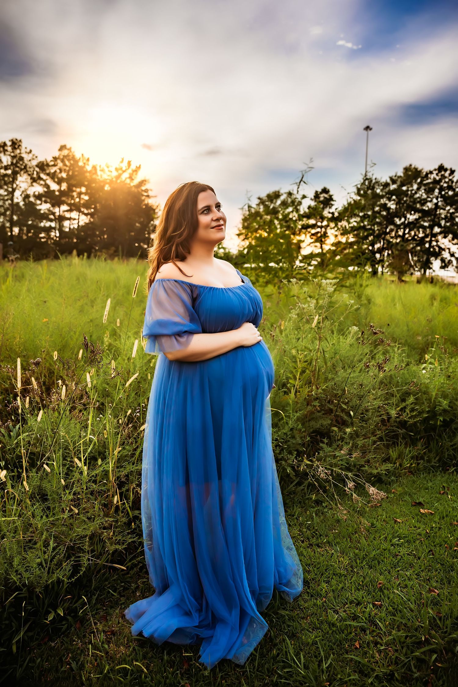 A woman in a flowing blue gown poses in a grassy field during golden hour.