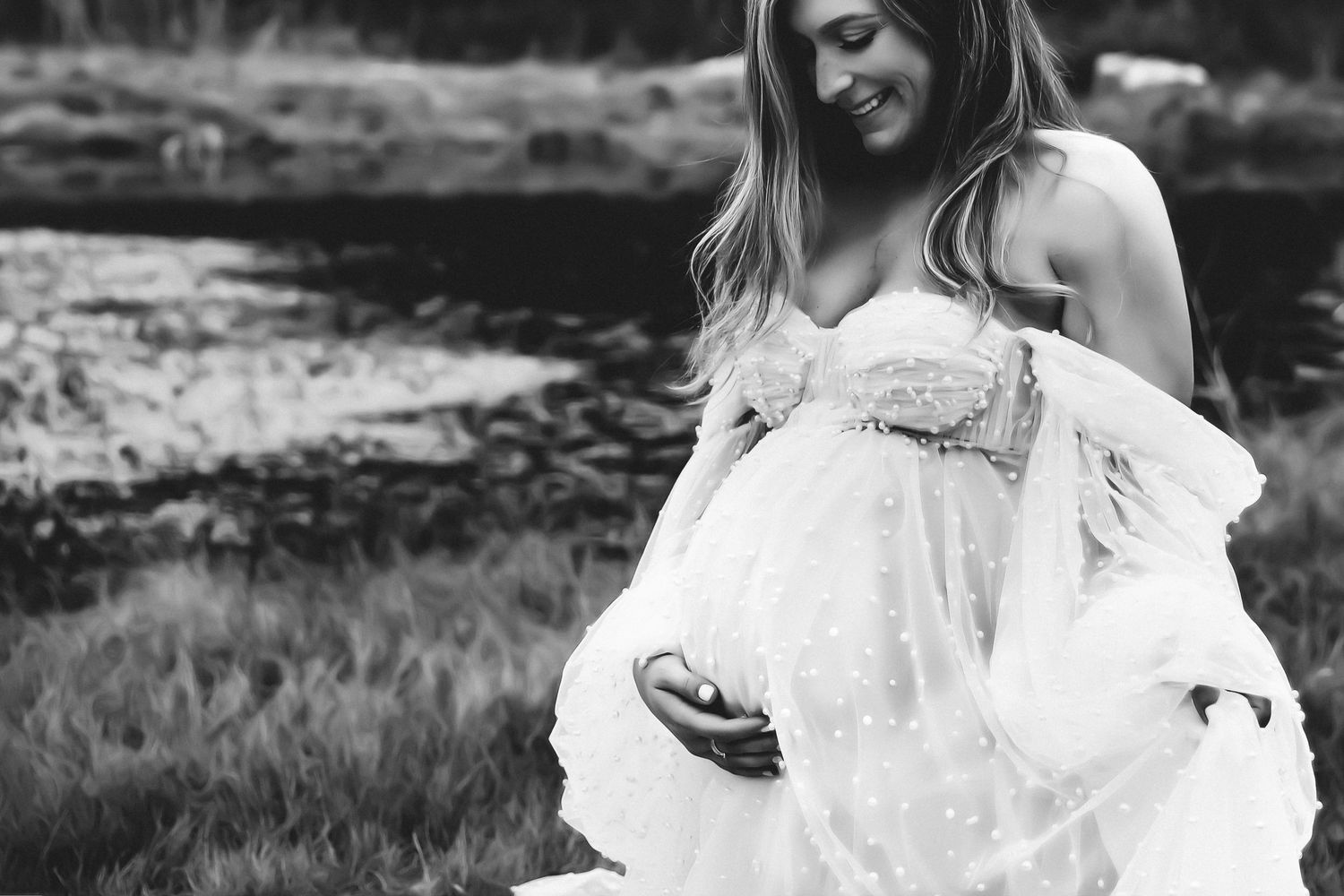 Dreamy black and white maternity photo of a woman in flowing white dress by a lake.