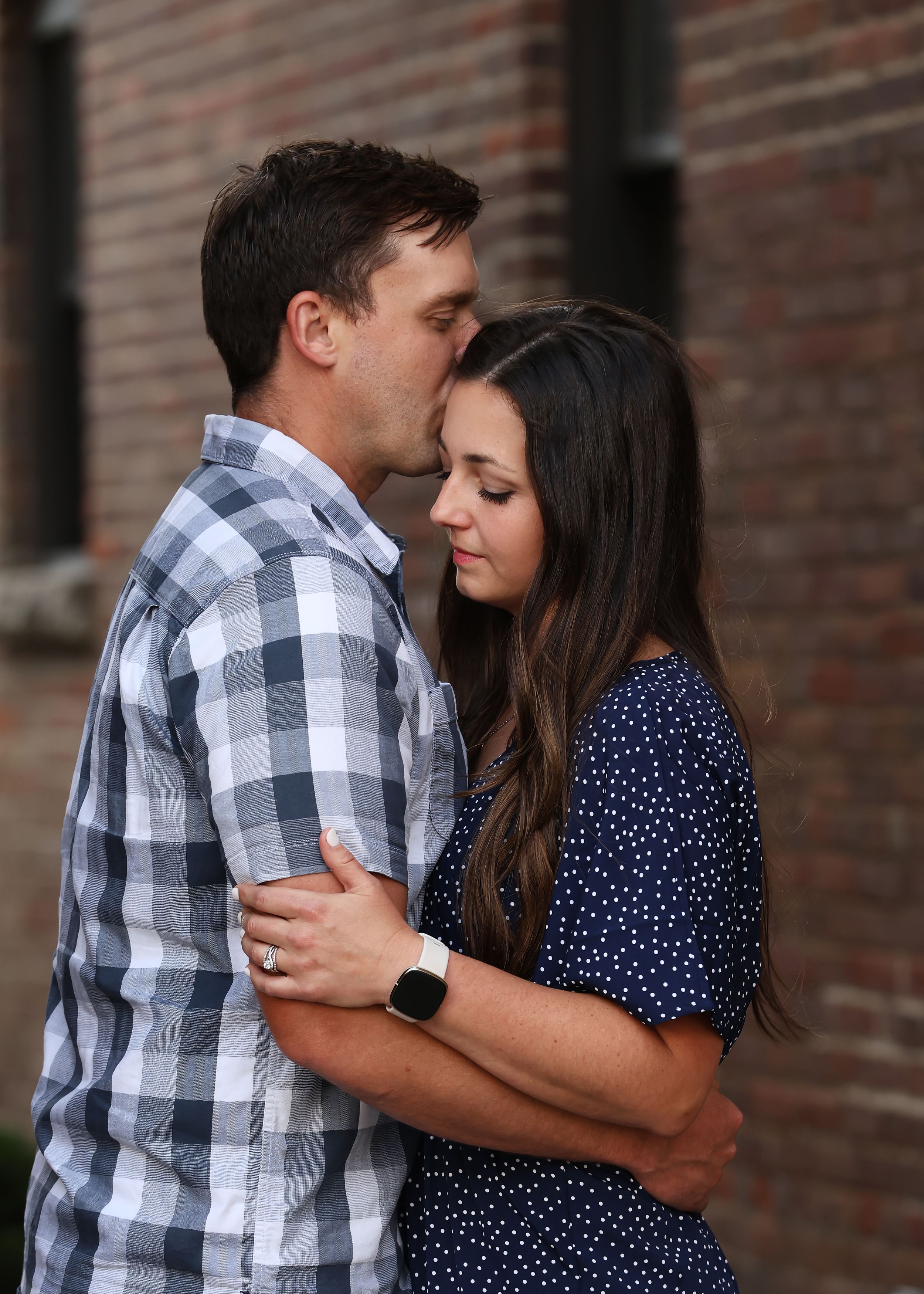 Couple embracing in front of brick wall wearing plaid and polka dot outfits.