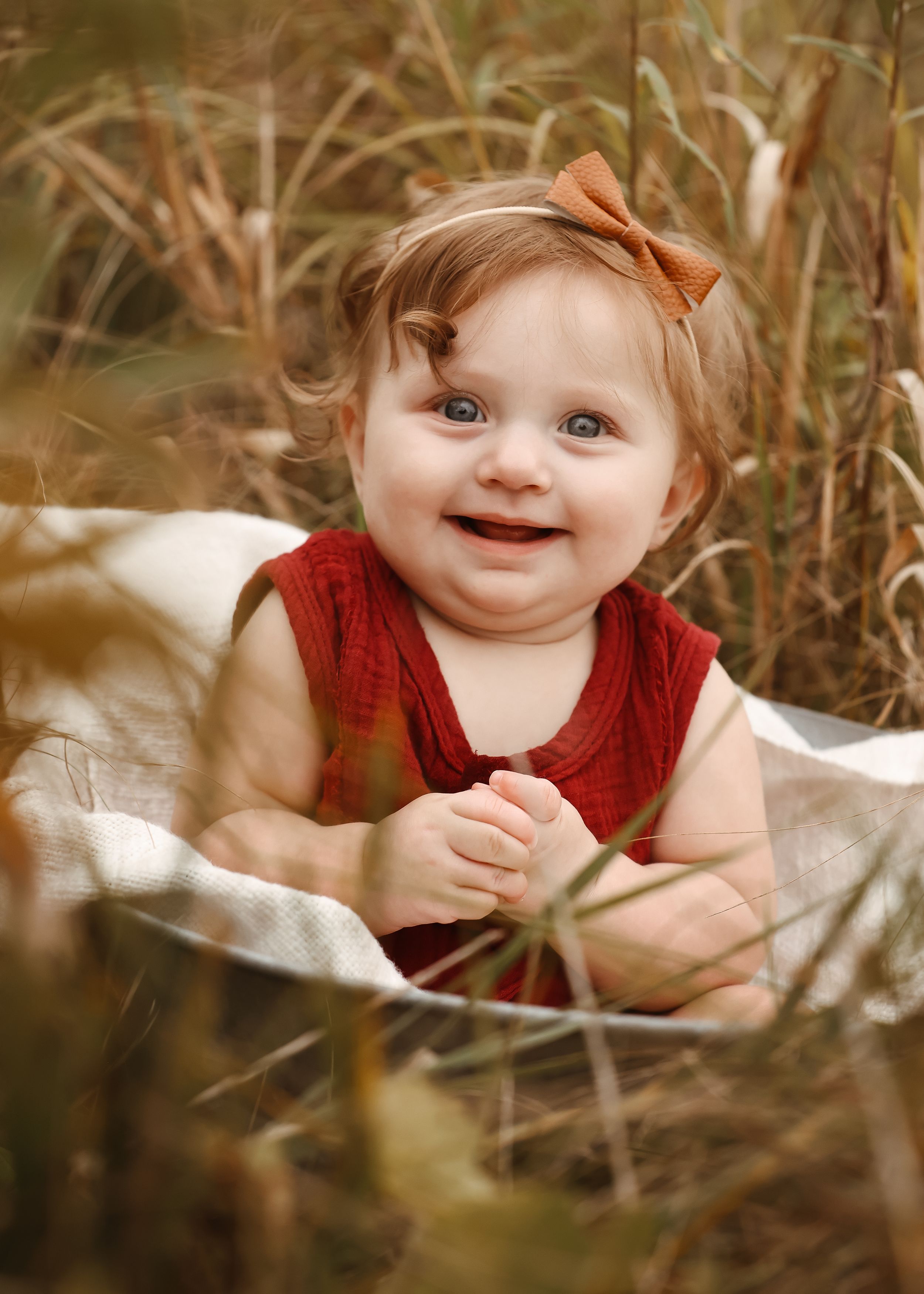 Baby in a red dress smiles joyfully while sitting among tall grass in a natural outdoor setting.