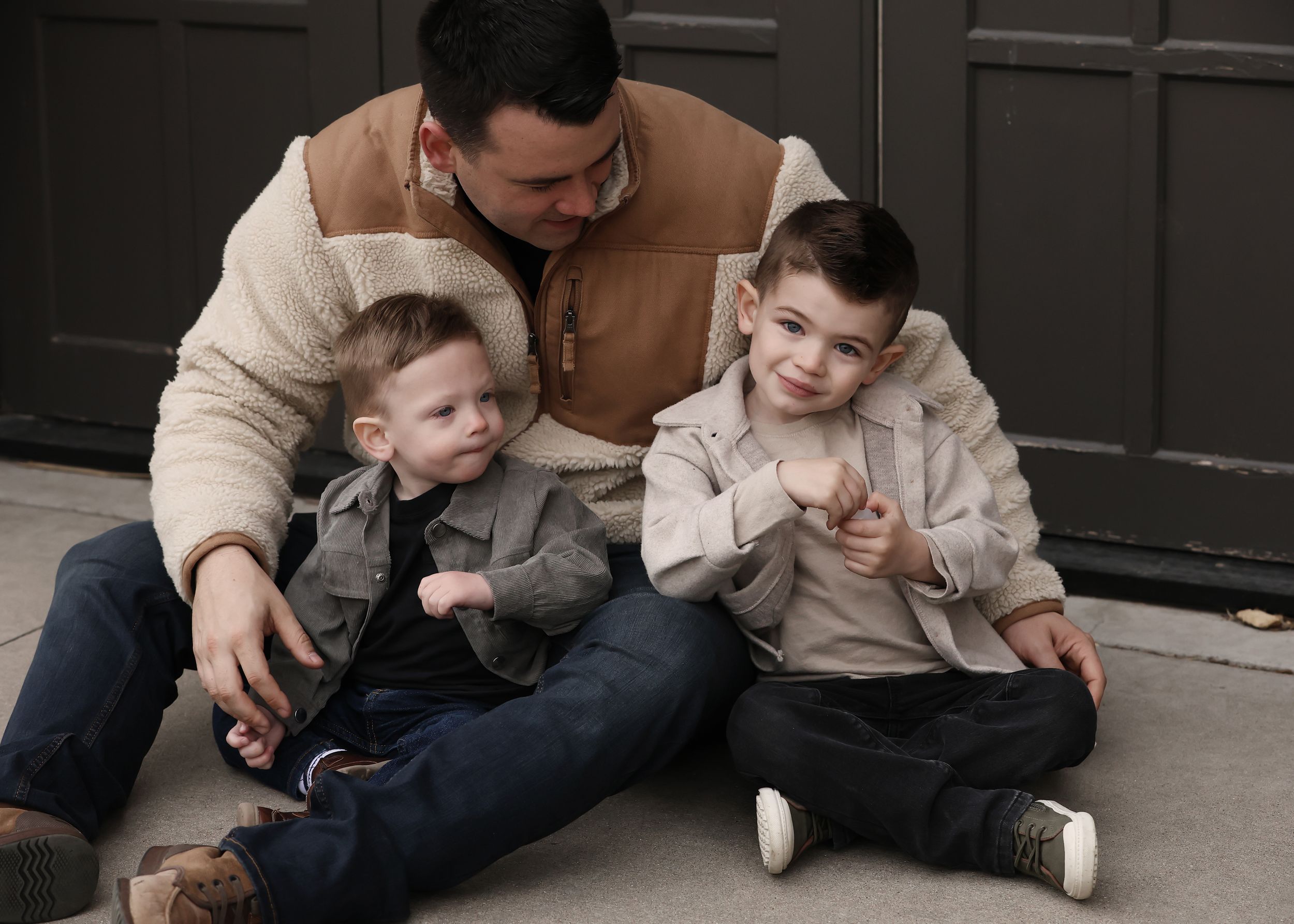 Family sitting together on floor wearing coordinated neutral tones against dark backdrop.