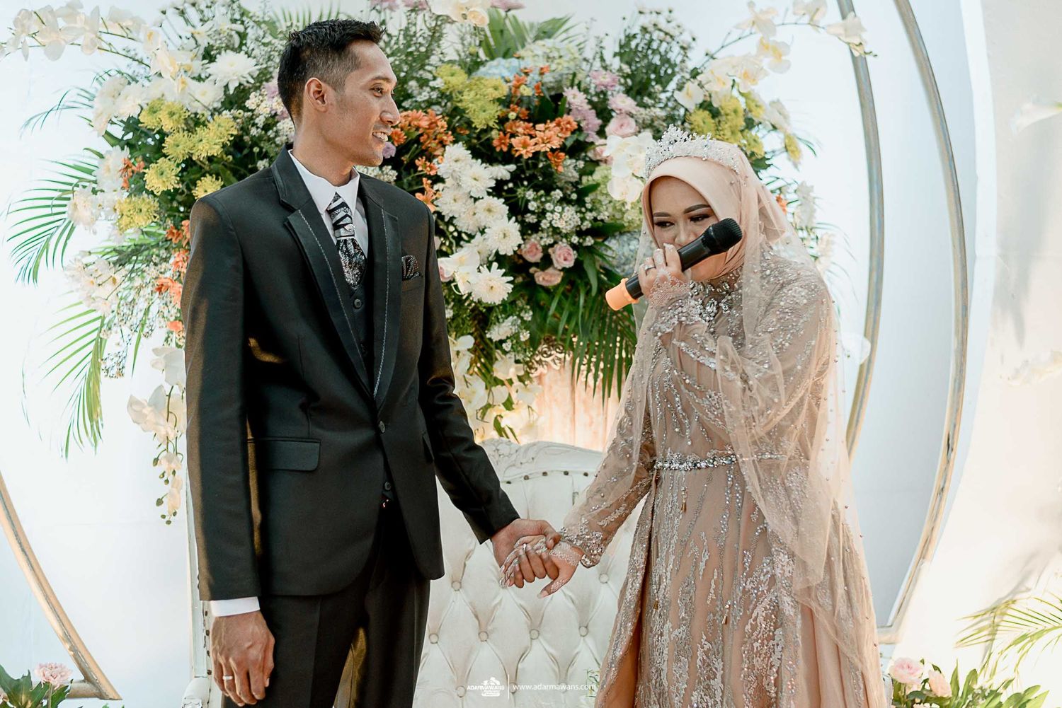 Joyful wedding couple holding hands under a decorated arch with yellow and white flowers.