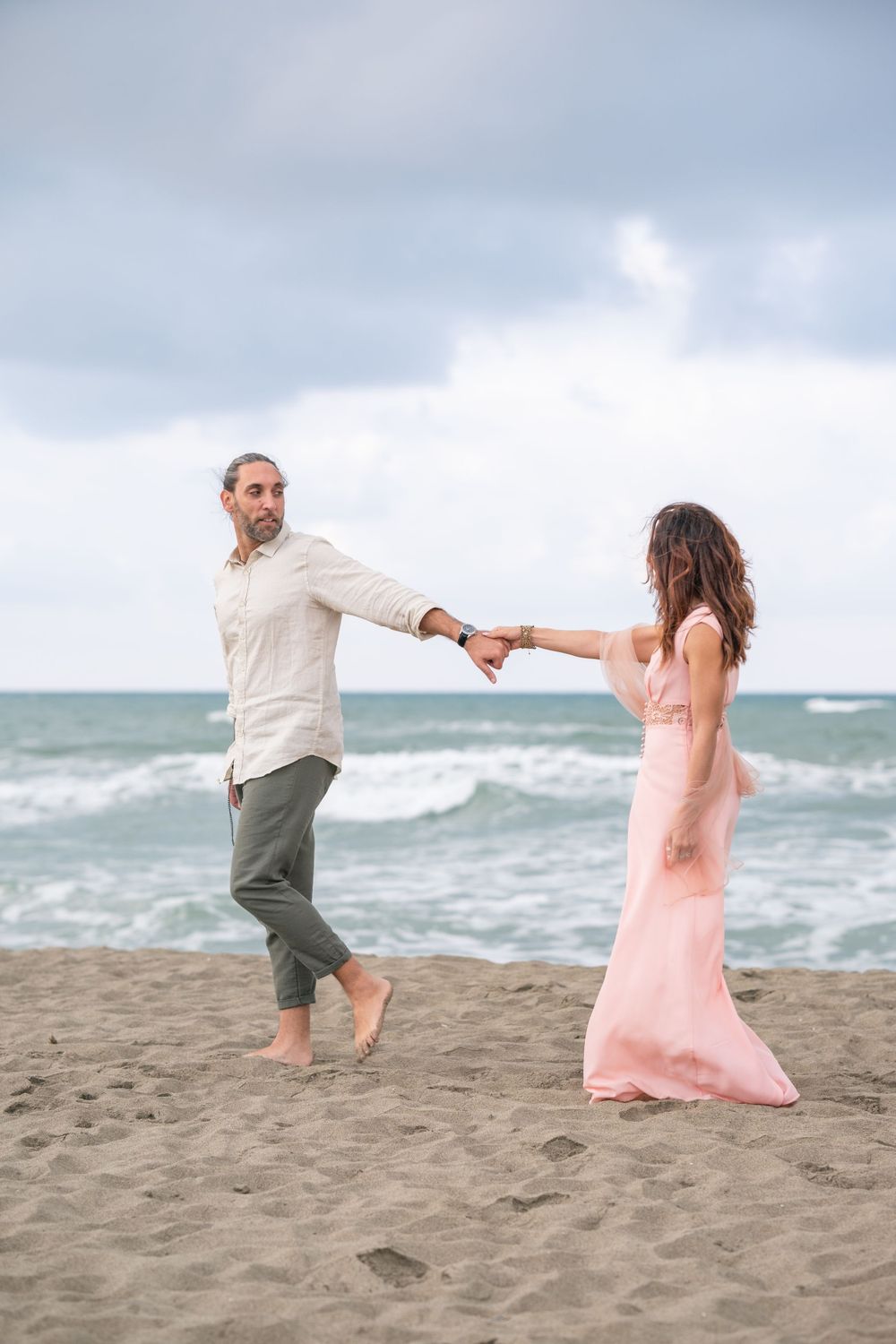 Couple dancing playfully on a beach with waves and cloudy sky in the background.