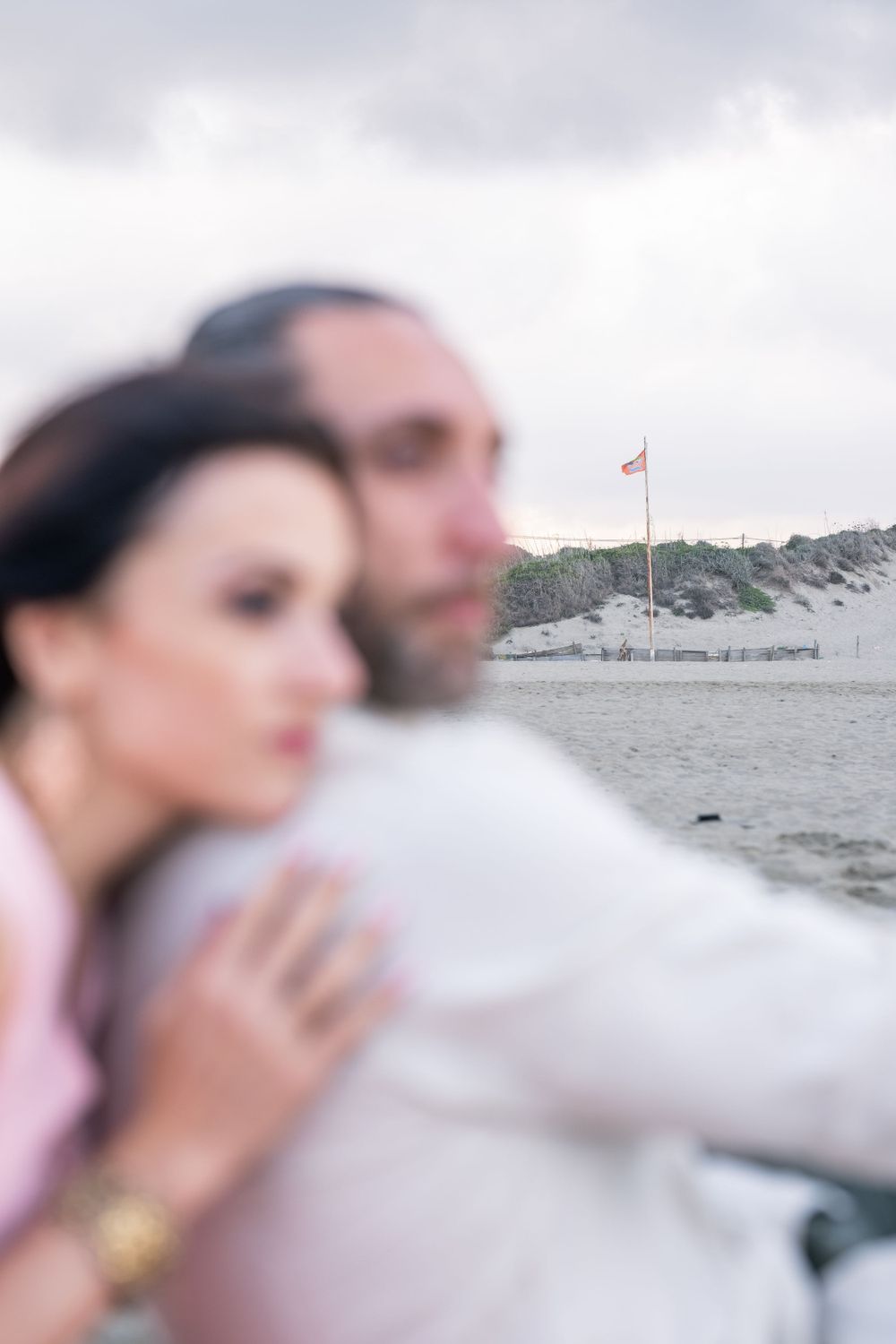 A couple embracing near the ocean with a flag pole visible in the background.