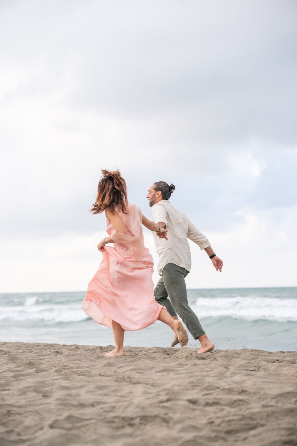 Couple joyfully dancing on beach with flowing pink dress catching the ocean breeze.