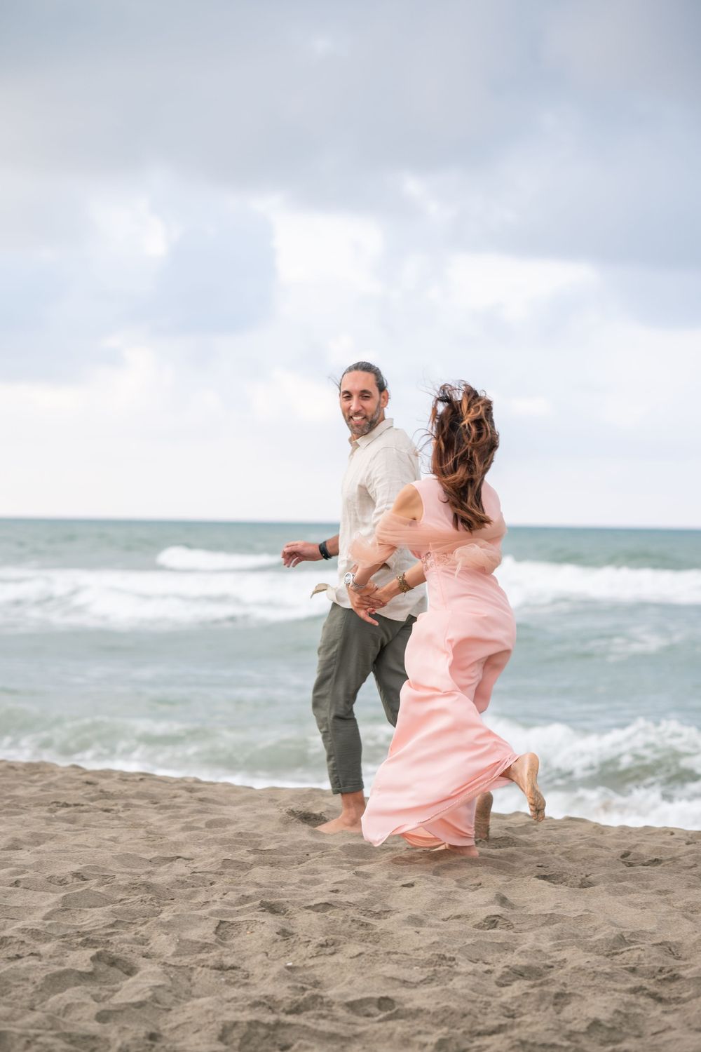 Romantic couple running together on sandy beach wearing pink dress and white shirt.