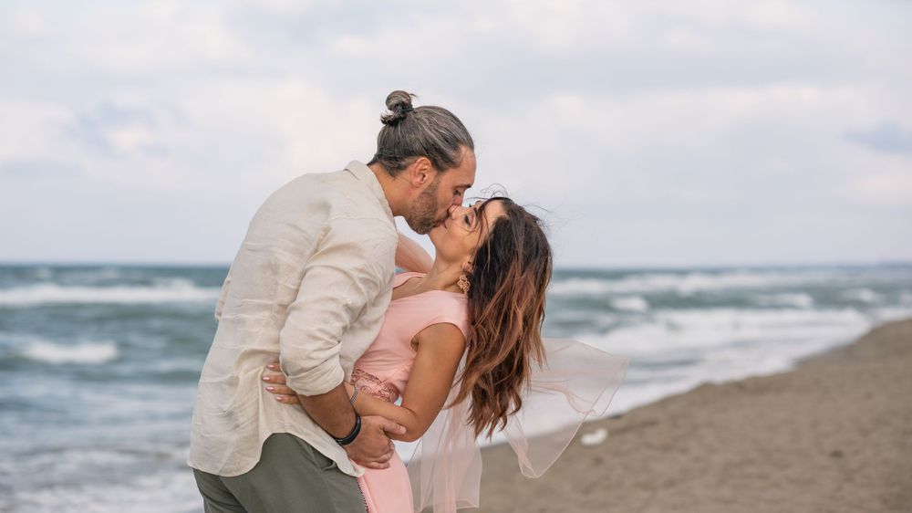 Intimate embrace on a windy beach with waves crashing behind.