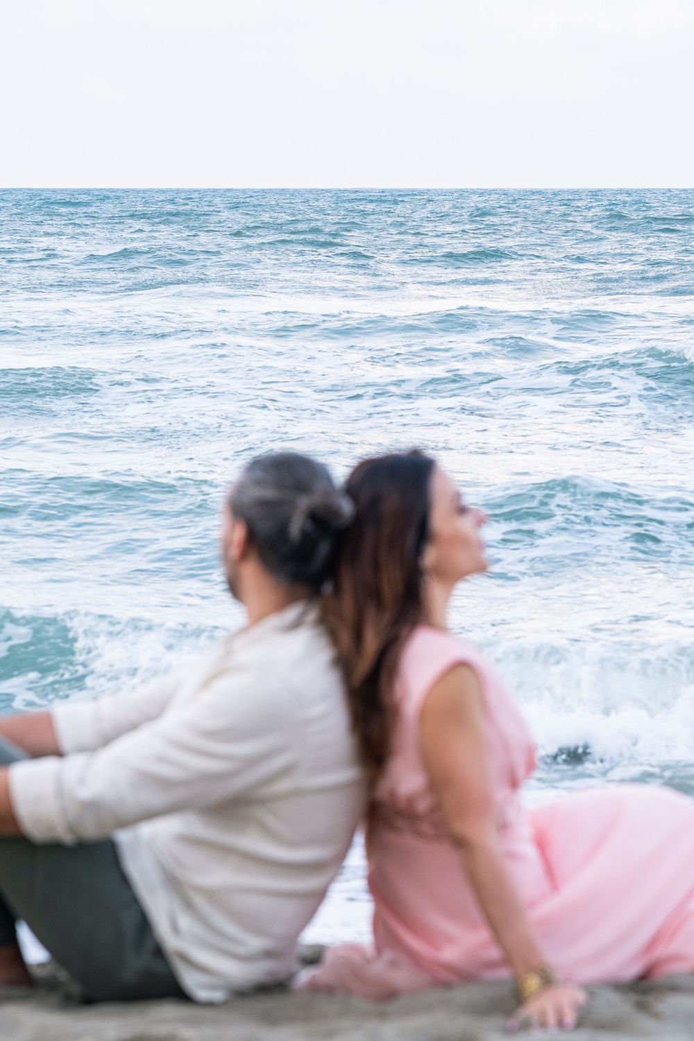 A couple sitting back-to-back on the beach watching the ocean waves.