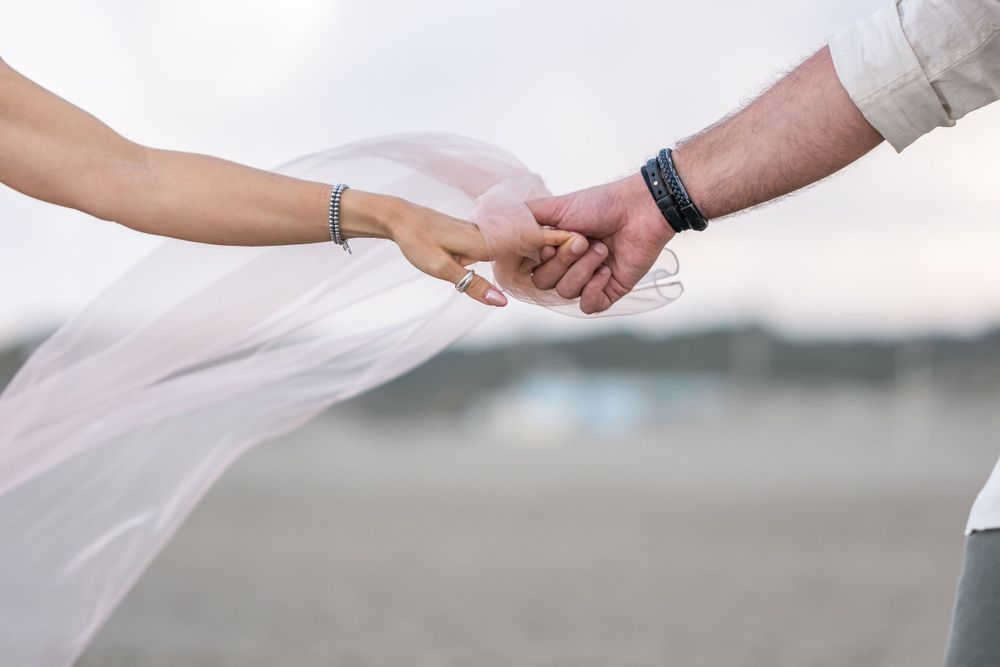 Close up of intertwined hands with a delicate veil flowing in the breeze on a beach.