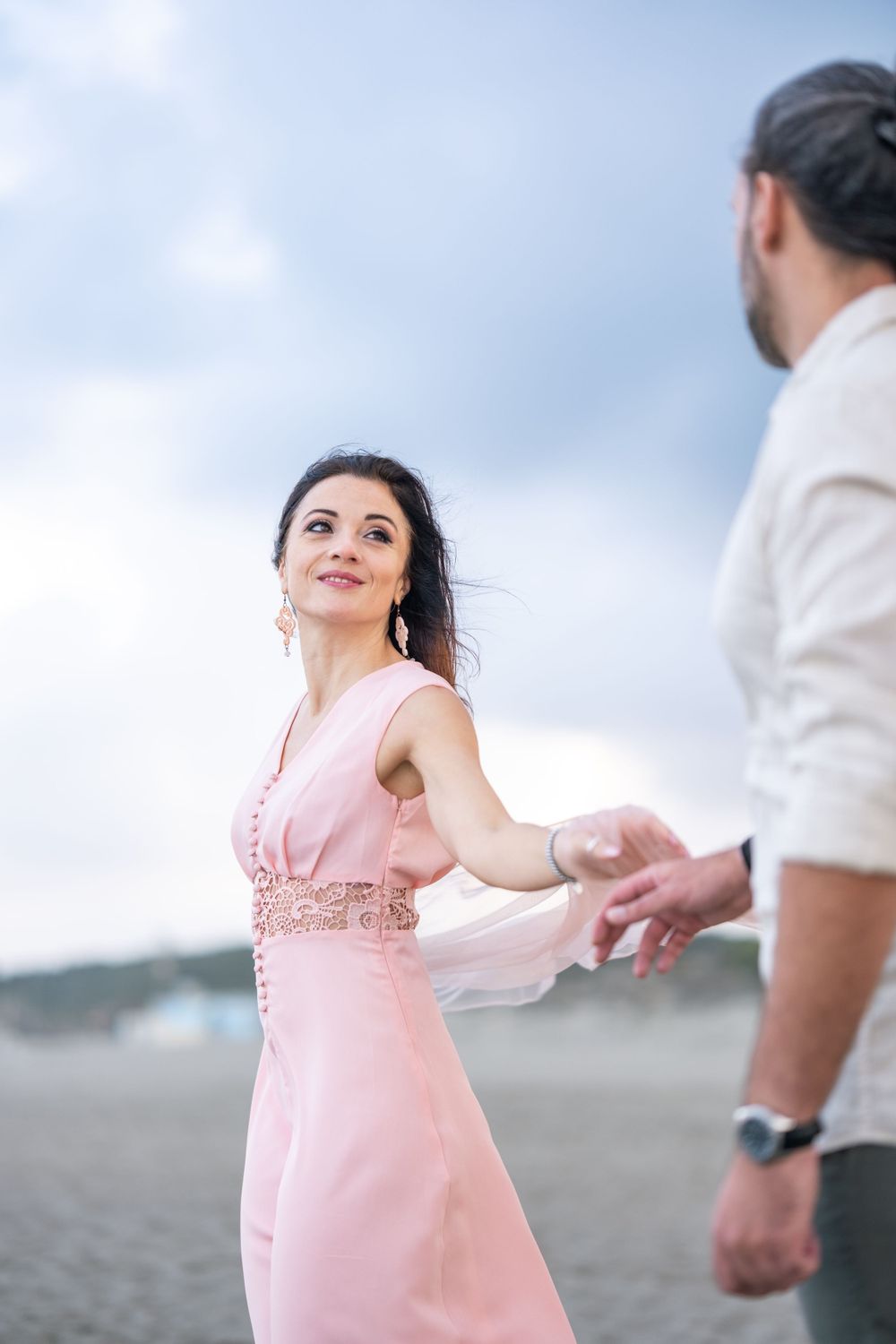 A couple dancing on the beach with the woman in a flowing pink dress against a cloudy sky.