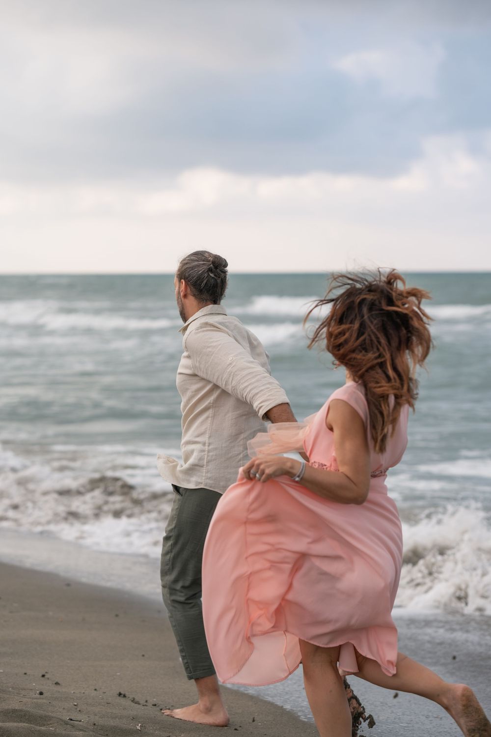 Couple dancing energetically on beach shore with dress flowing in wind.