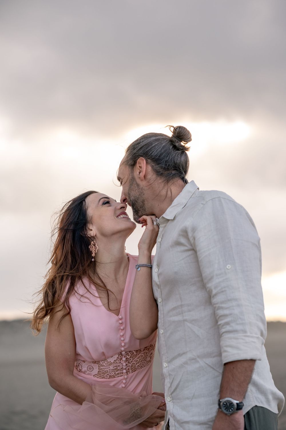 A romantic couple sharing an intimate moment at sunset on a beach, with the woman wearing a pink dress.
