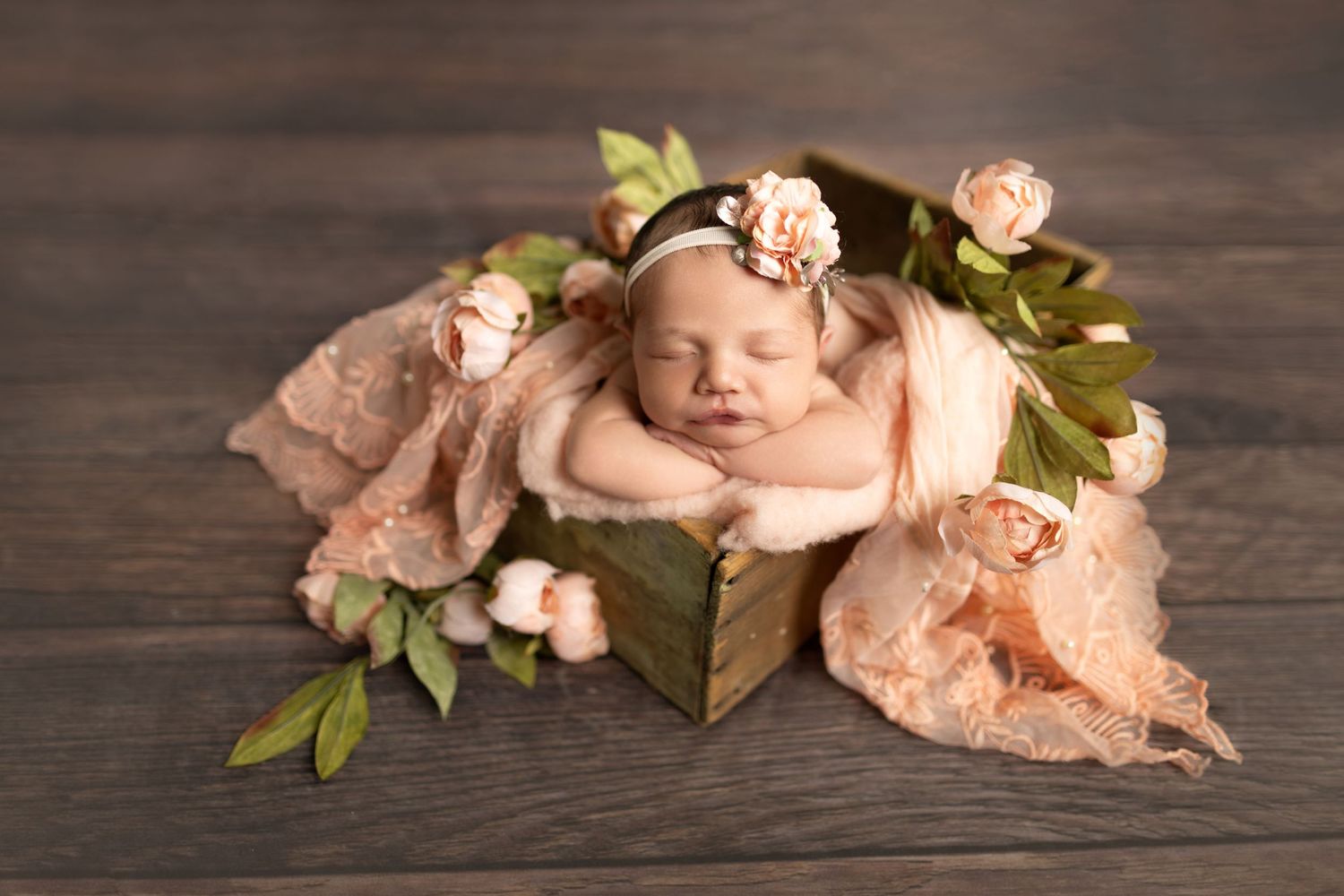 Sleeping newborn posed with peach fabric and floral decorations on wooden background.