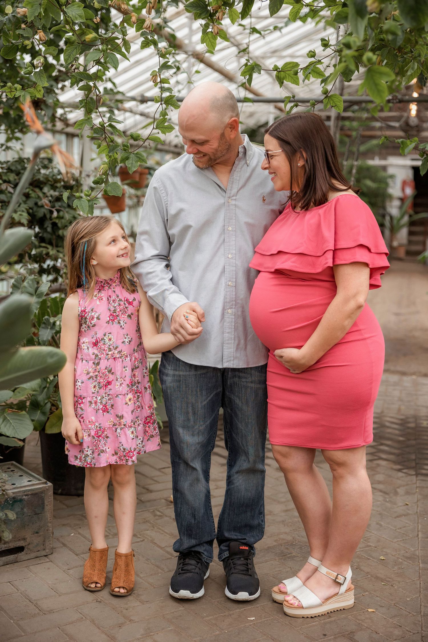 An expectant family shares a tender moment together in a plant-filled greenhouse setting.