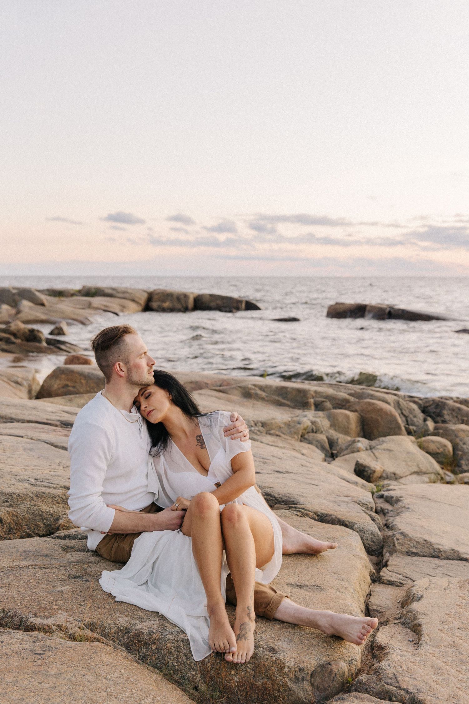 Romantic couple in white clothing sitting on coastal rocks at sunset.